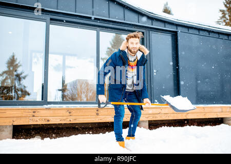 Schöner mann im winter Kleidung Schnee Reinigung mit einer Schaufel in der Nähe der modernen Haus in den Bergen Stockfoto