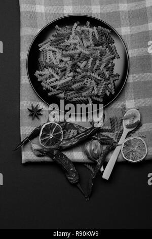 Leckere bunte Italienische Pasta in Keramik Teller mit Löffel aus Holz, Chili, Orange, Knoblauch, Sternanis und Zimt weiß und blau karierten Stoff oder karierten Tuch auf dunklem Hintergrund, Ansicht von oben Stockfoto