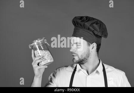 Koch in Burgund einheitliche hält Glas mit Grütze. Frühstück und gesundes Essen Konzept. Kochen mit nachdenklichen Gesicht und jar. Mann mit Bart hält Glas Glas mit Porridge auf rotem Hintergrund. Stockfoto