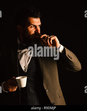 Business- und fünf Minuten Pause Konzept. Kerl in klassischen Anzug hat Kaffeepause. Mann mit Bart hält Zigarre auf schwarzem Hintergrund. Geschäftsmann mit nachdenklichen Gesicht Getränke Tasse heiße Getränke- und raucht. Stockfoto