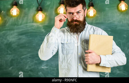 Zurück zum Konzept der Schule. Lehrer in Brillen ab Lektion. Bärtige hipster hält Buch, schwarzes Brett für den Hintergrund. Mann mit Bart und Schnurrbart auf ernstes Gesicht vor der Tafel. Stockfoto