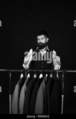 Mann mit Bart in Weste von Wäscheständer. Designer in der Nähe von Kleiderbügeln. Mode und individuellen Stil. Maßgeschneiderte mit ernsten Gesicht hält Maßband in der Nähe von benutzerdefinierten Jacken auf schwarzem Hintergrund Stockfoto