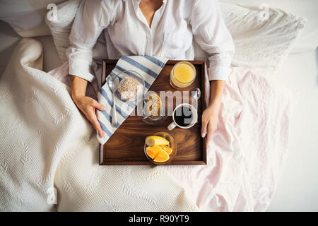 Ein Mittelteil der Frau im Bett lag, mit Kaffee und Frühstück drinnen in der Morgen in einem Schlafzimmer, mit Smartphone. Eine Ansicht von oben. Stockfoto