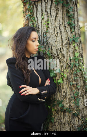 Schöne junge Frau mit verschränkten Armen lehnte sich gegen einen Baum Stockfoto