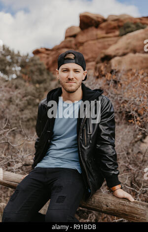 Porträt der Jungen gut aussehenden Casual Reisen schöner Mann außerhalb lächelnd in der Nähe von Ancient Desert Red Rocks in Jacke Stockfoto