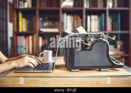 Frau Hände Typ auf modernen Notebook. Alte vintage Staubbedeckte Schreibmaschine mit Blatt weißes Papier in der Nähe von modernes Notebook auf Bücherregal Hintergrund. Moderne Stockfoto