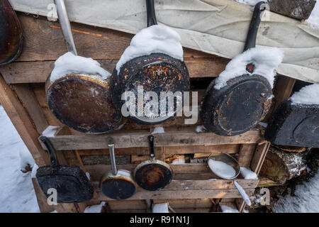 Alte Pfannen in verbrannte Asche hängen an der Wand. Eine warme Suche Ferienhaus in der Mitte von einem verschneiten Winterwald. Brennholz in Stapeln, mit Schnee bedeckt. geschnitten Trunks in einer Hütte im Winter Wald Stockfoto