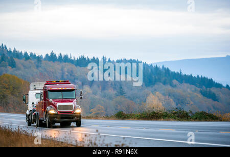 Big Rig kraftvolle Rot American Motorhaube semi Abschleppwagen mit Scheinwerfer abschleppen gebrochen Semi Truck mit Dachspoiler auf der kurvenreichen Abend twilig gedreht Stockfoto