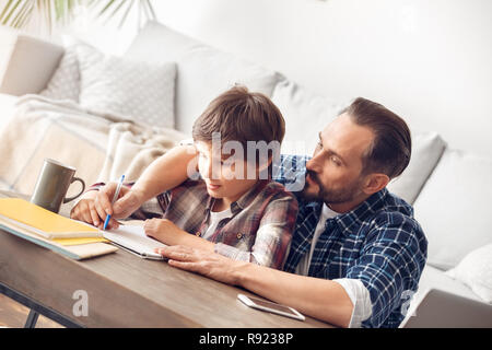 Vater und Sohn zu Hause am Tisch Vater umarmt Jungen seine Hand Abtretung schriftlich im Notebook Holding sitzt konzentriert Stockfoto