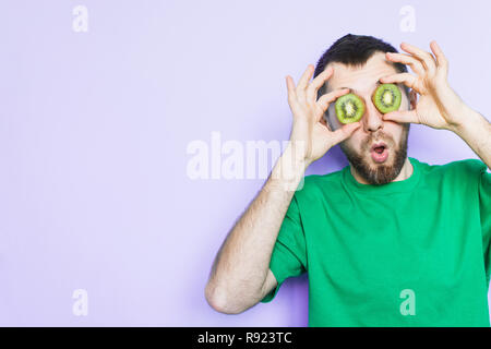 Junge bärtige Mann mit Scheiben von grüne Kiwi vor seinen Augen, überrascht und schockiert. Hellpurpurroten Hintergrund, kopieren. Stockfoto