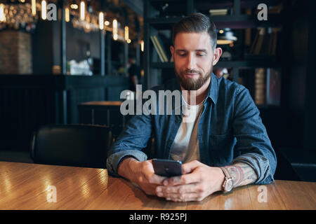 Junge Bartgeier Geschäftsmann, in einem denim Hemd bekleidet, am Tisch im Cafe und Verwendung des Smartphones zu sitzen. Mann mit Gadget. Stockfoto