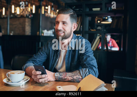 Junge Bartgeier Geschäftsmann, in einem denim Hemd bekleidet, am Tisch im Cafe und Verwendung des Smartphones zu sitzen. Mann mit Gadget. Stockfoto