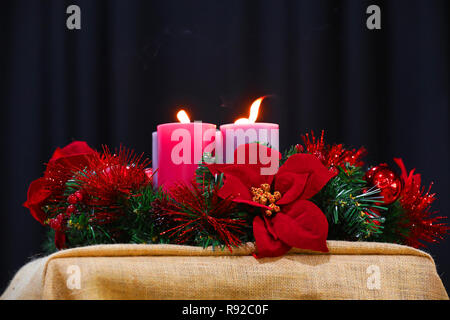 Eine rote und grüne Imitation Christmas wreath Dekoration mit Lametta und Weihnachtsstern Blumen umkreisen große Kerzen und Flammen. Xmas-Konzept Stockfoto