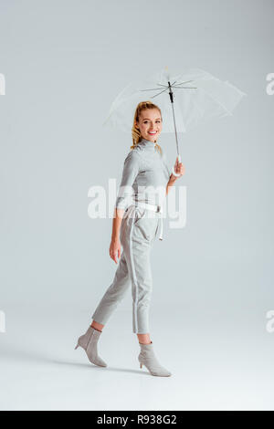 Frau in grauer Kleidung an Kamera und mit transparenten Regenschirm auf grauem Hintergrund posiert Stockfoto