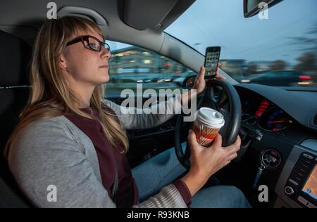 Junge Frau Auto fahren in die Innenstadt, während Ihr Handy und eine Tasse Kaffee gleichzeitig Holding Stockfoto