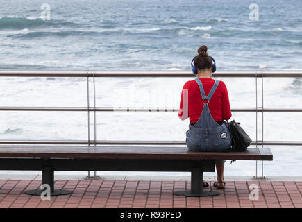 Ansicht der Rückseite des junge Frau mit Kopfhörern auf Sitz mit Blick auf das Meer. Stockfoto