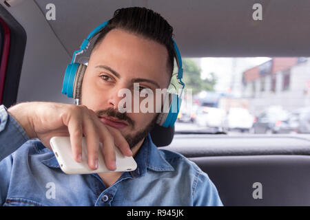 Junge, urbane, Geschäftsmann professionelle tragen Jeanshemd auf der Rückbank des Autos, das Hören von Musik auf einem Smartphone. Konzept der Bestrebungen, Konzentration Stockfoto