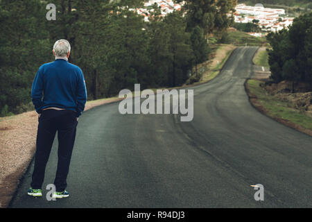 Active Senior Mann stand auf der einsamen Straße zwischen Bergen. Ältere Menschen rücken auf einsamen Highway Stockfoto