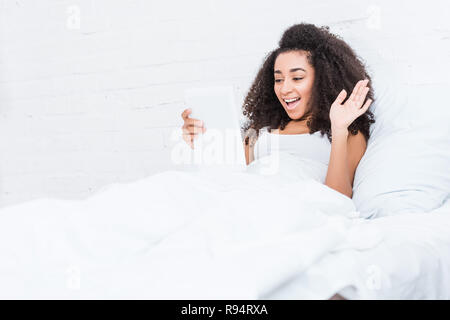 Lachend afrikanische amerikanische Frau, video Anruf auf digital Tablet und winkte mit der Hand im Bett zu Hause. Stockfoto