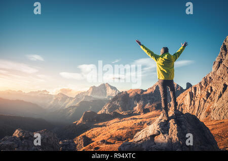 Sportliche Mann stand auf dem Stein mit erhobenen Armen gegen die Berge bei Sonnenuntergang. Glücklich, junger Mann, Felsen, Wald und blauem Himmel in Dolomit Stockfoto