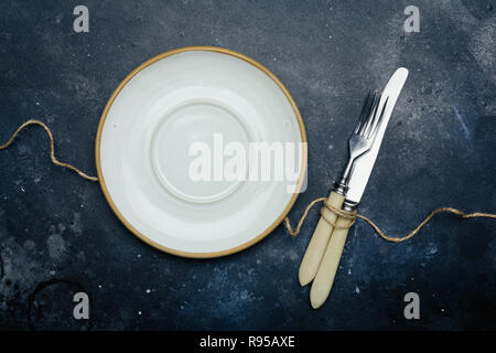 Leere weiße Platte (Keramik, Rustikal) auf einem dunkelblauen Hintergrund mit Messer und Gabel. Kopieren Sie Platz. Stockfoto