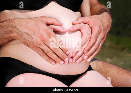 Herzförmige Schwangerschaft. Hände von zukünftigen Eltern das Herzsymbol auf schwangeren Bauch. Ziemlich schwangere Frau Hug mit ihrem Ehemann. Warten des Babys. Stockfoto