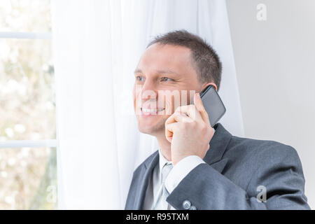 Nahaufnahme der Junge, glücklich, Lächeln, Mann, männlich Geschäftsmann in grauen Anzug, Krawatte, Bräutigam, durch die Fenster in der Wohnung, Haus, Zimmer, weiße Vorhänge Stockfoto