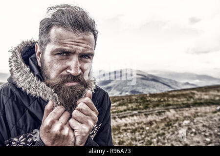 Mann. Kalt. bärtiger Mann, langen Bart, brutale kaukasischen Hipster mit Schnurrbart auf ernstes Gesicht. Unrasierten Kerl mit stilvollen Haare, bärte Haarschnitt kauern von kalt auf dem Berg oben an bewölkten Himmel. Stockfoto