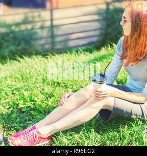 Schöne rothaarige Mädchen sitzen auf dem Gras und trinken Kaffee aus einem Pappbecher. Lifestyle Frühstück im Park Stockfoto