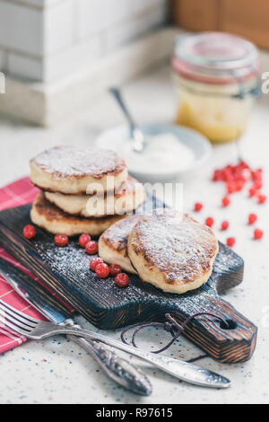 Auf einem weißen Tisch aus Kunststein ist ein Holz Schneidebrett mit fünf Quark Pfannkuchen und rote Beeren. Stockfoto