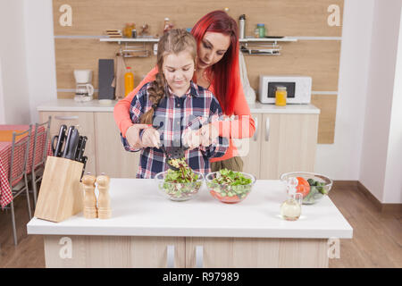 Lächelnde Mutter mischen Salat Zutaten mit ihrer Tochter helfen. Die Mumie wenig helfen. Stockfoto