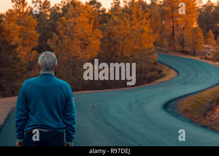 Aktiver älterer Mann steht alleine auf einsamen Straße zwischen Bergen. Ältere Mann zu Fuß zurück auf einsamen Highway Stockfoto