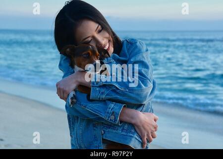 Eine junge Frau, die ihrem kleinen Hund umarmt am Strand bei Dämmerung. Laguna Beach, Kalifornien. Stockfoto
