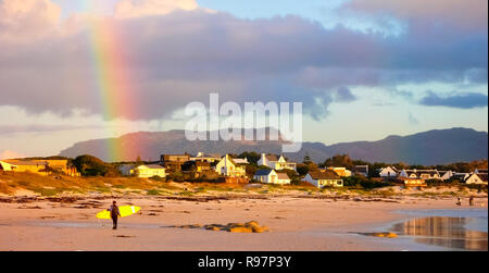 Ein junger Surfer bei Sonnenuntergang am langen Strand von Noordhoek Kapstadt mit einem Regenbogen im Hintergrund Stockfoto