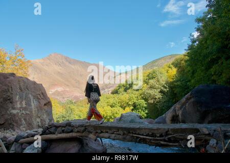 Eine weibliche Touristen überquert eine Brücke über einen Bach in den Ausläufern des Atlasgebirges. Die bewaldeten Imlil Tal und die Berge sind hell erleuchtet in der Stockfoto