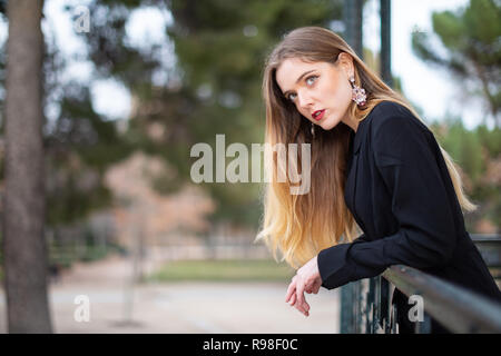 Seitenansicht der attraktive junge Frau im eleganten Outfit lehnte sich auf Zaun und weg schauen beim Stehen auf unscharfen Hintergrund der Park Stockfoto