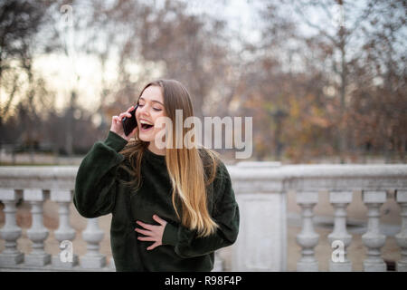 Hübsche junge Dame vor Lachen, laut, während im Herbst Park stehen und in Smartphone Gespräch Stockfoto