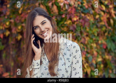 Lächelnden jungen Frau Plaudern auf einem mobilen im Herbst vor einem bunten Kriechgang glücklich in die Kamera lächeln mit Sun flare Stockfoto