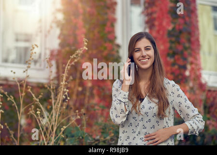 Lächelnden jungen Frau Plaudern auf einem mobilen im Herbst vor dem Weißen Haus mit bunten Kriechgang glücklich lächelnd in die Kamera mit Sun fla Stockfoto