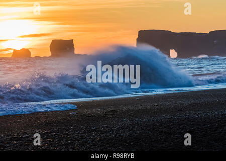 Wellen, die am Sonnenuntergang mit Dyrhólaey, ein Felsen mit einem Bogen, der in der Entfernung entlang der schwarze Sandstrand in der Nähe des Dorfes Vik im Winter in Island Stockfoto