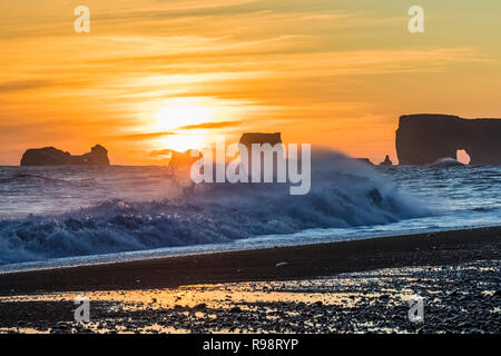 Wellen, die am Sonnenuntergang mit Dyrhólaey, ein Felsen mit einem Bogen, der in der Entfernung entlang der schwarze Sandstrand in der Nähe des Dorfes Vik im Winter in Island Stockfoto