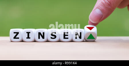 Hand dreht einen Würfel und verändert die Richtung, in die sich ein Pfeil symbolisiert, die Zinssätze ("Zinsen" in Deutsch) nach oben (oder umgekehrt) Stockfoto