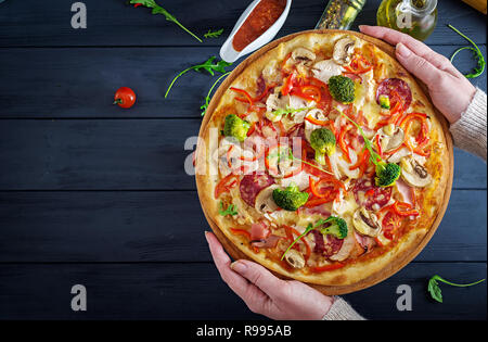 Frische italienische Pizza mit hühnerfilet, Champignons, Schinken, Salami, Tomaten, Käse auf einem schwarzen Hintergrund in den Händen. Italienisches Essen. Ansicht von oben Stockfoto