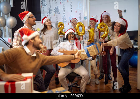 Lächelnd business Arbeiter haben Spaß und Tanz in Santa hat bei der Weihnachtsfeier und Geschenke auszutauschen im Büro Stockfoto