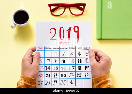 Hohe Betrachtungswinkel von einem kaukasischen Mann beobachten ein Kalender von 2019 an einem gelben Tisch sitzen neben einer Tasse Kaffee, eine Brille und eine grüne Keine Stockfoto