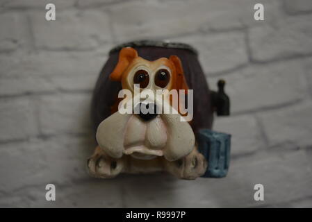 Keramische Abtropfschale in Form eines Bierfasses mit einem Hundekopf und einem blauen Glas Bier daneben vor dem Hintergrund einer weißen Ziegelwand. Stockfoto