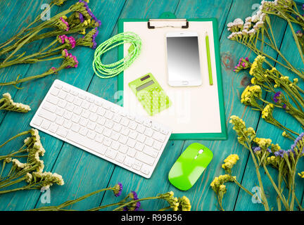 Arbeitsplatz mit wireless Slim Keyboard, Maus, Smart Phone, Zwischenablage, Rechner, leeres Papier, Bleistift, Netzkabel, bunte Blumen auf blau Holz des Stockfoto