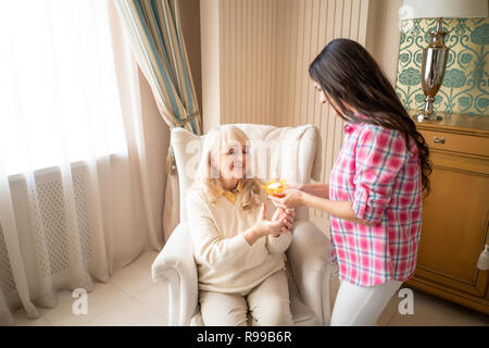 Schöne ältere Mutter nimmt Ihre Kleinen Geburtstag Kuchen mit einer brennenden Kerze aus ihrer süßen Tochter. Feier. Indoor, Stockfoto