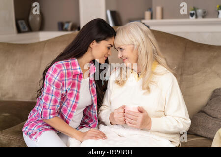 Lächelnde Mutter hält eine Tasse Kaffee beim Sitzen auf der Couch und Berührung der Stirn mit Ihrer schönen Tochter. Stockfoto