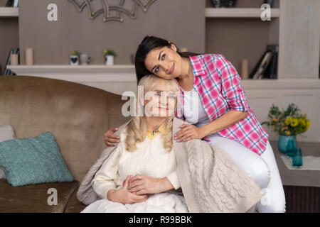 Charmante Portrait von älteren Mutter und liebende Tochter. Reife Frau sitzt auf der Couch mit Ihrer Tochter umarmte Ihre. Stockfoto